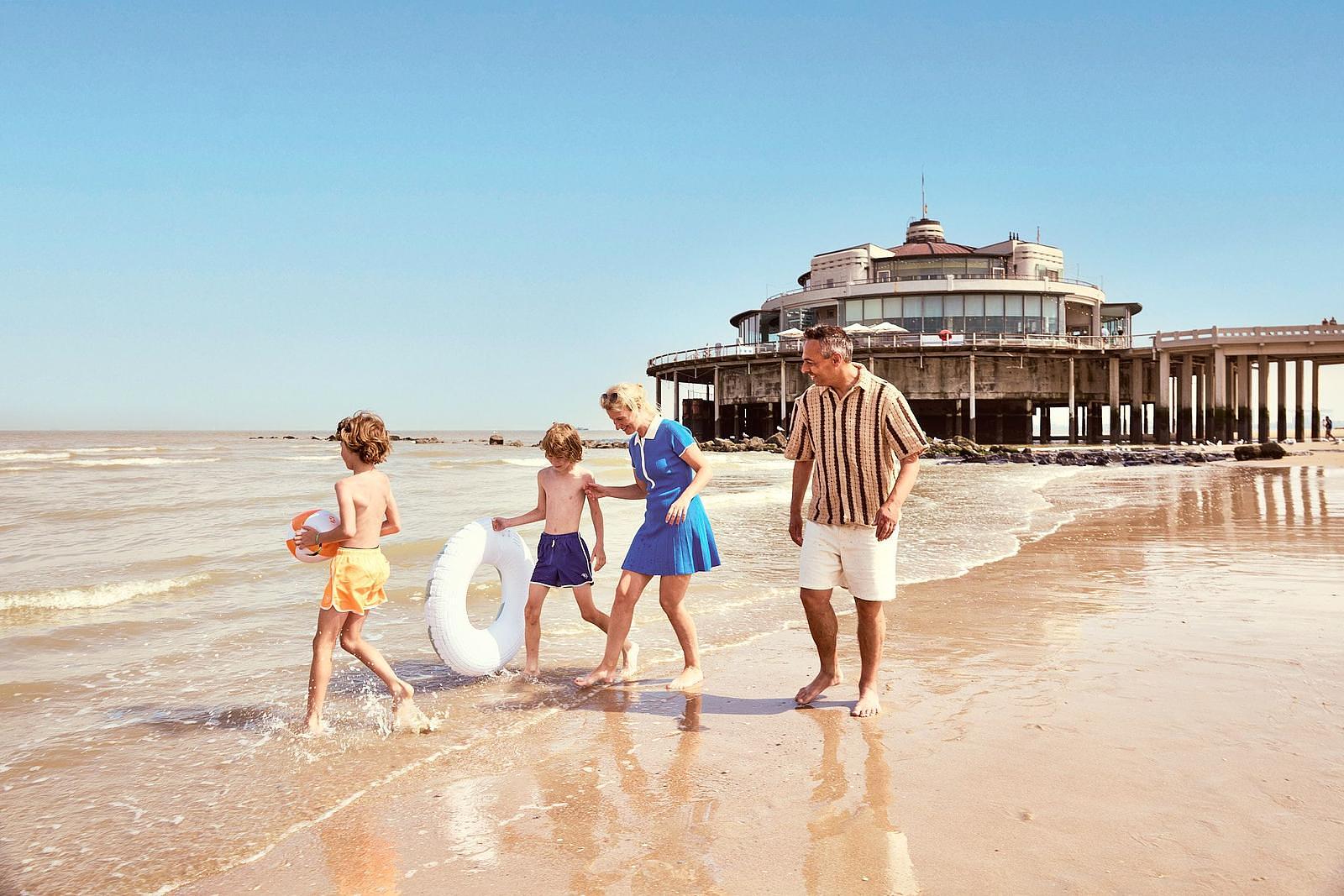 Een vrouw en een man wandelen met hun kinderen langs de waterlijn op het strand in Blankenberge, terwijl de kinderen spelen met een bal en een zwemband onder een heldere blauwe lucht.