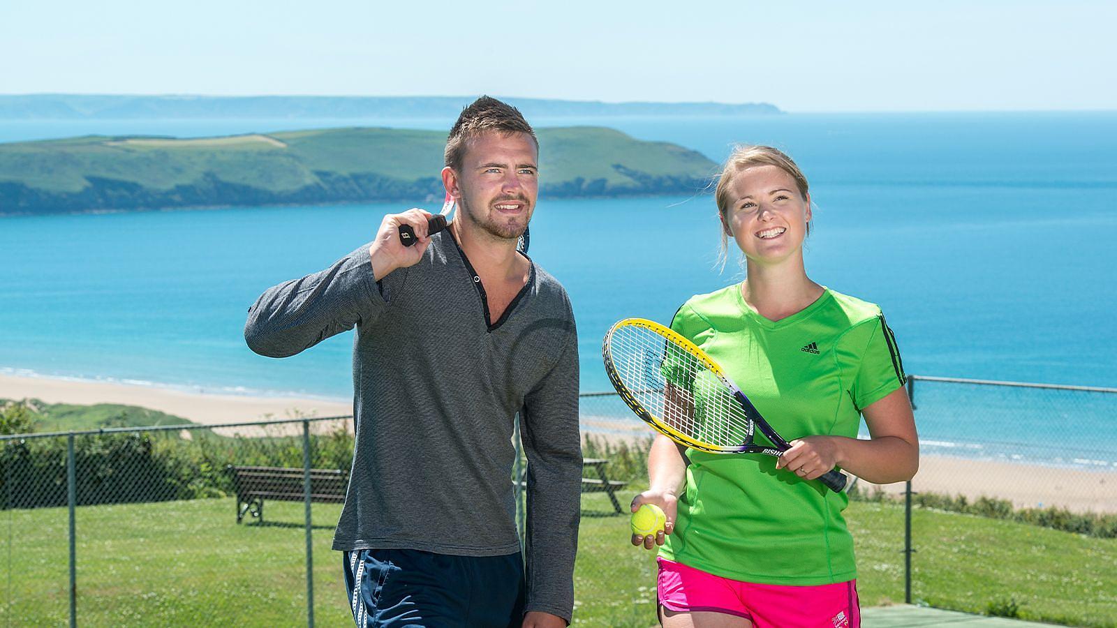 A couple holding tennis racquets and ball walking from the tennis court at Woolacombe Bay Holiday Park with the ocean behind.