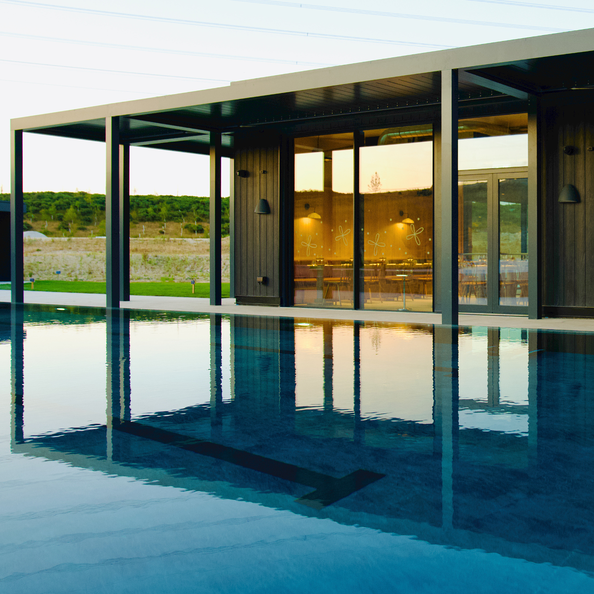 Firefly Spa's 25m heated outdoor swimming pool at Silverlake Estate, Dorset, with calm blue water reflecting the Firefly Restaurant.